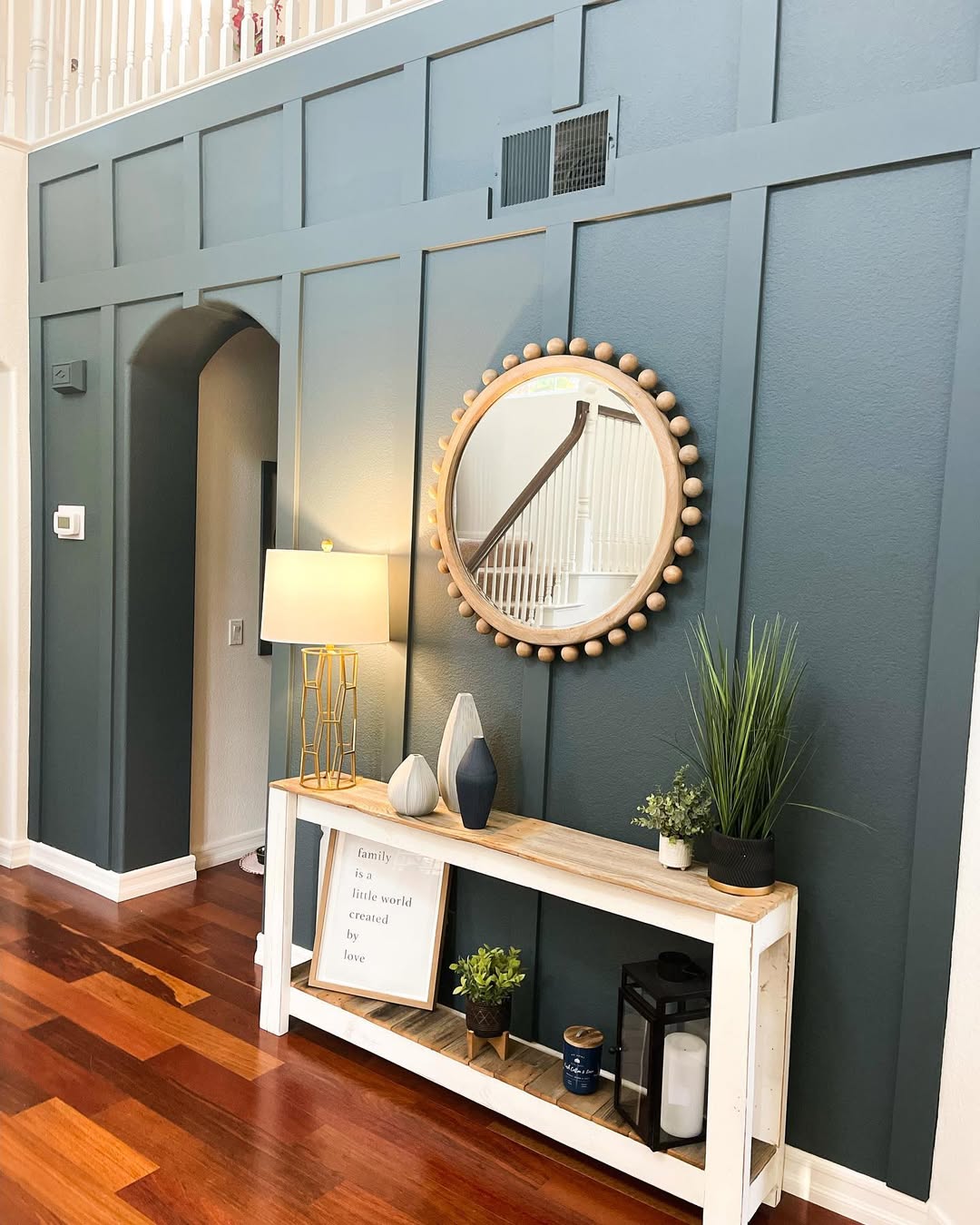 A modern entryway with a blue-green wood accent wall, round mirror, table lamp, decorative vases, plants, and a white console table on wood flooring. Perfect for those seeking fresh accent wall ideas.