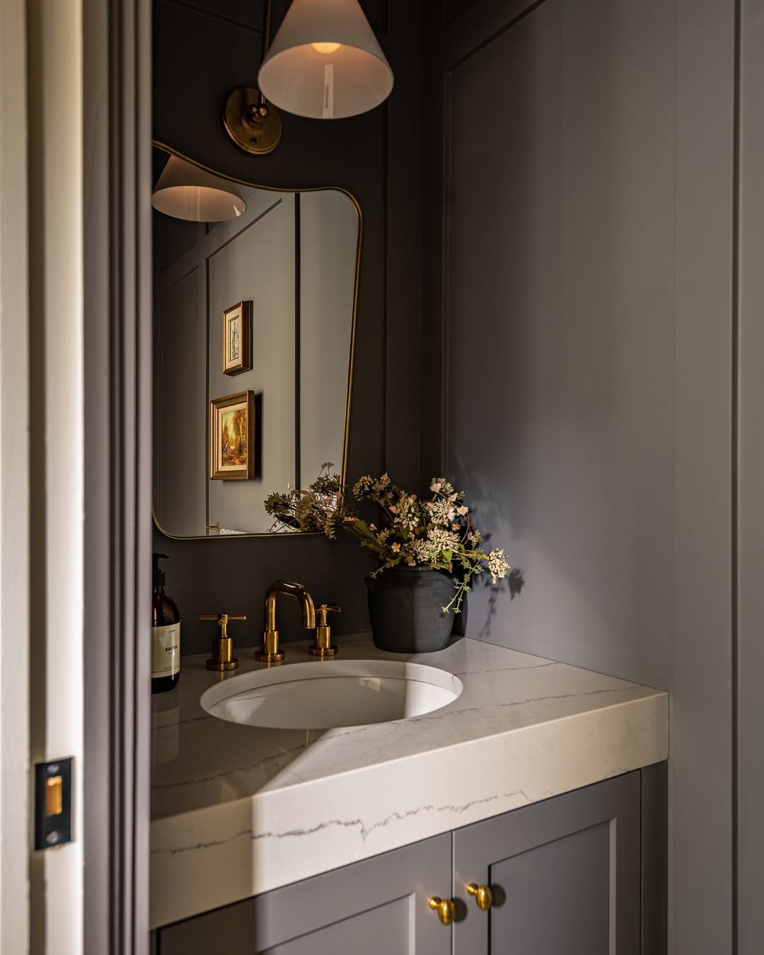 A small bathroom vanity with a marble countertop, brass fixtures, a black vase with flowers, a mirror, and framed art on dark paneled walls creates one of those stylish powder rooms with true wow-factor decor.
