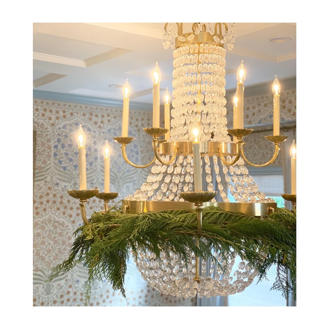 A crystal chandelier with lit candles is decorated with a ring of green foliage, offering festive Christmas chandelier decor as it hangs from the ceiling in a patterned room.