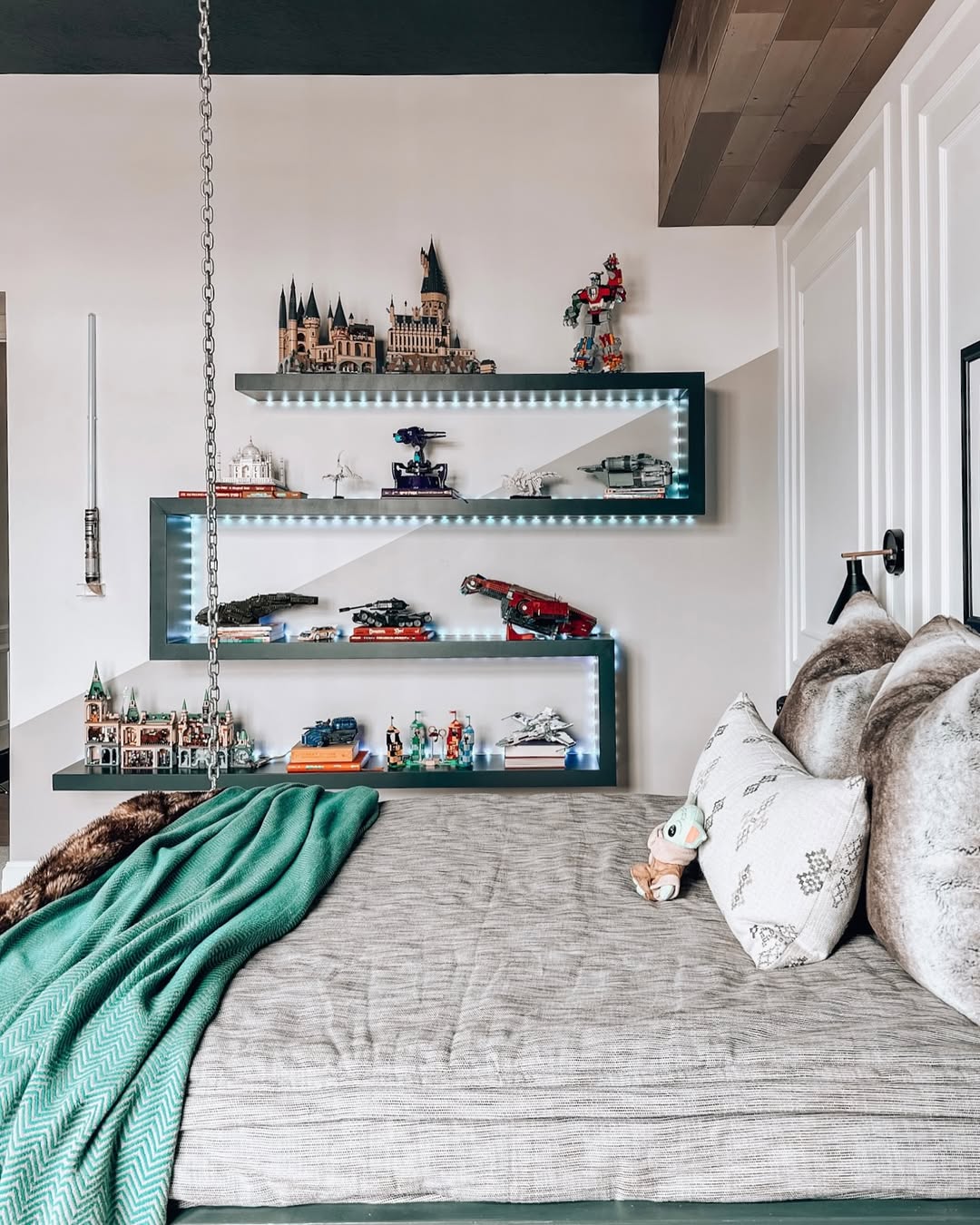 A neatly made bed with green blanket and pillows sits beside wall-mounted shelves displaying Lego models, including Harry Potter sets, under strip lighting—perfect for boys bedroom ideas and smart storage solutions.