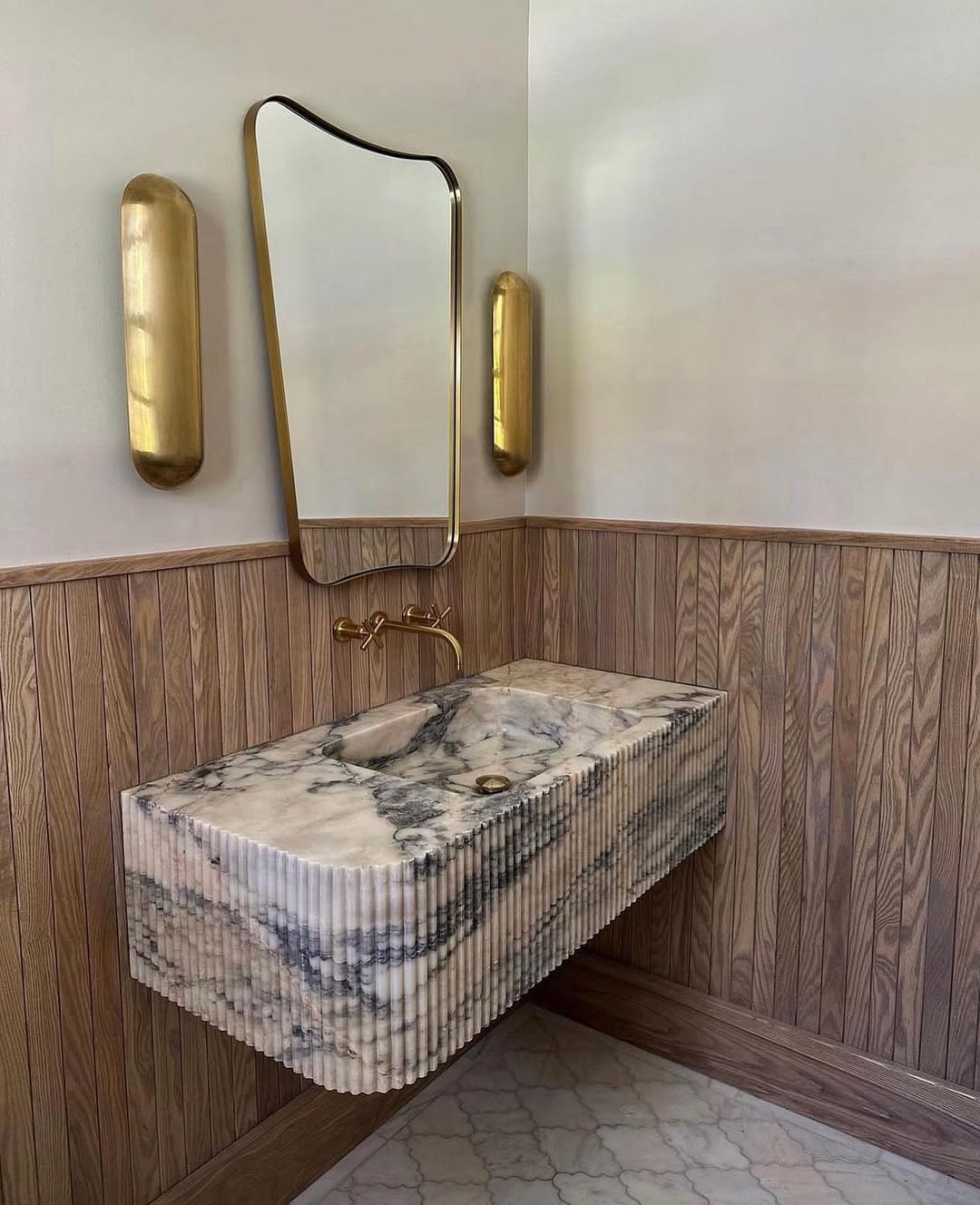 A marble sink with a fluted front, mounted on wood-paneled walls, with a gold-framed mirror and two gold wall sconces above—perfect for those seeking wow-factor powder rooms or stylish bathroom ideas.