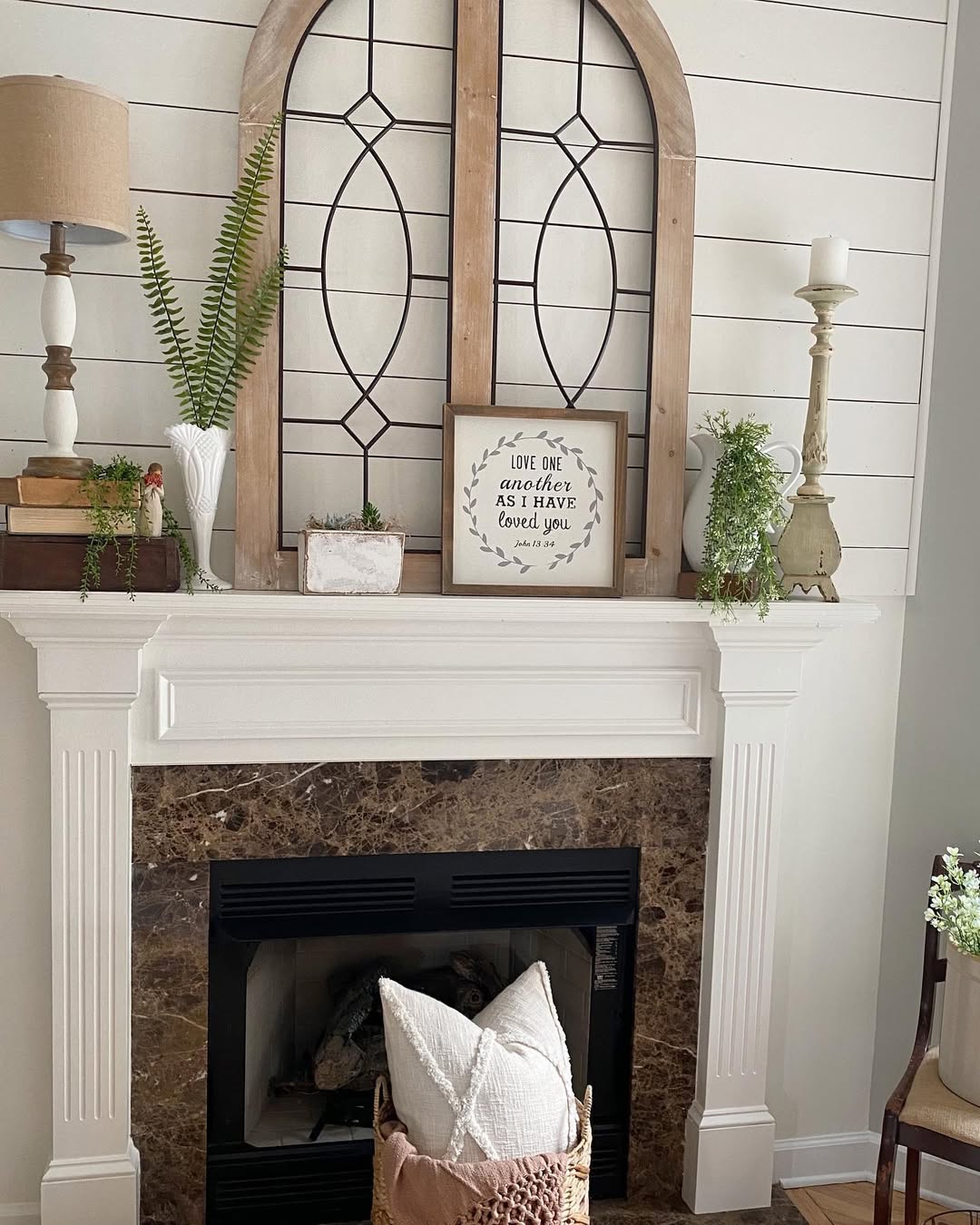 A white mantel with decorative items, including lamps, plants, books, a framed quote, and a wooden window frame, enhances the fireplace design above a brown marble fireplace.