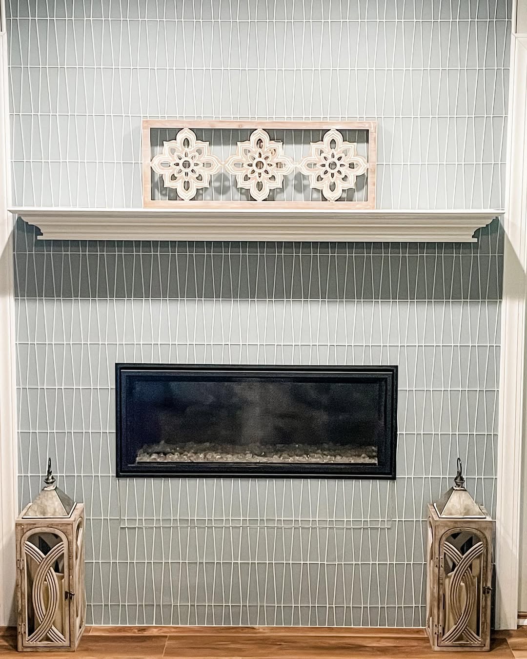 A modern fireplace with a black rectangular opening, flanked by decorative lanterns and a white panel on the shelf above. The wall showcases a geometric pattern, offering stylish fireplace tile ideas for contemporary spaces.