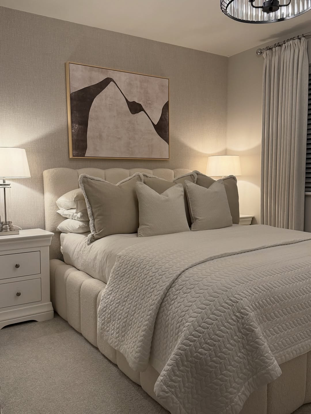 A neatly made bed with a beige quilt, multiple pillows, bedside tables with lamps, and neutral wall art offers stylish inspiration for those seeking cozy guest bedroom ideas in a softly lit space.
