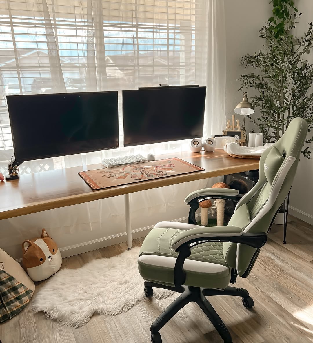 A modern home office with dual monitors, keyboard, mouse on a large desk showcases smart desk organization, featuring an ergonomic chair, white rug, potted plant, lamp, and plush dog pillow by a window with sheer curtains.