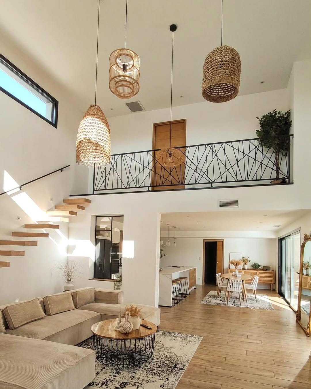 Modern open-plan living room and kitchen with high ceilings, floating staircase, beige furniture, woven pendant lights, and large windows letting in natural light—perfect for those seeking high ceiling living room ideas.