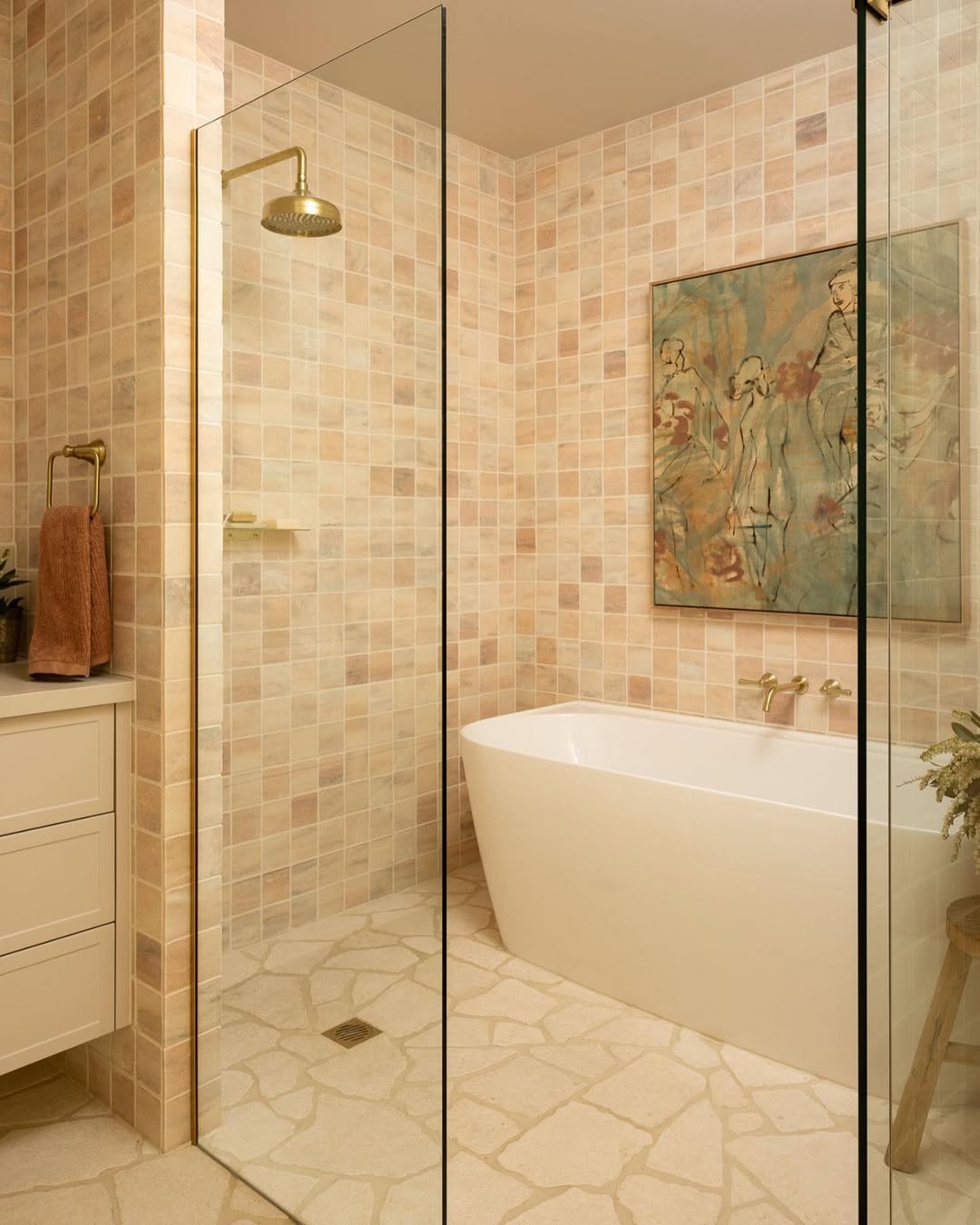 Modern small bathroom with beige tile walls, a freestanding white bathtub, framed artwork above the tub, and a stylish walk-in shower with a brass showerhead and no door.