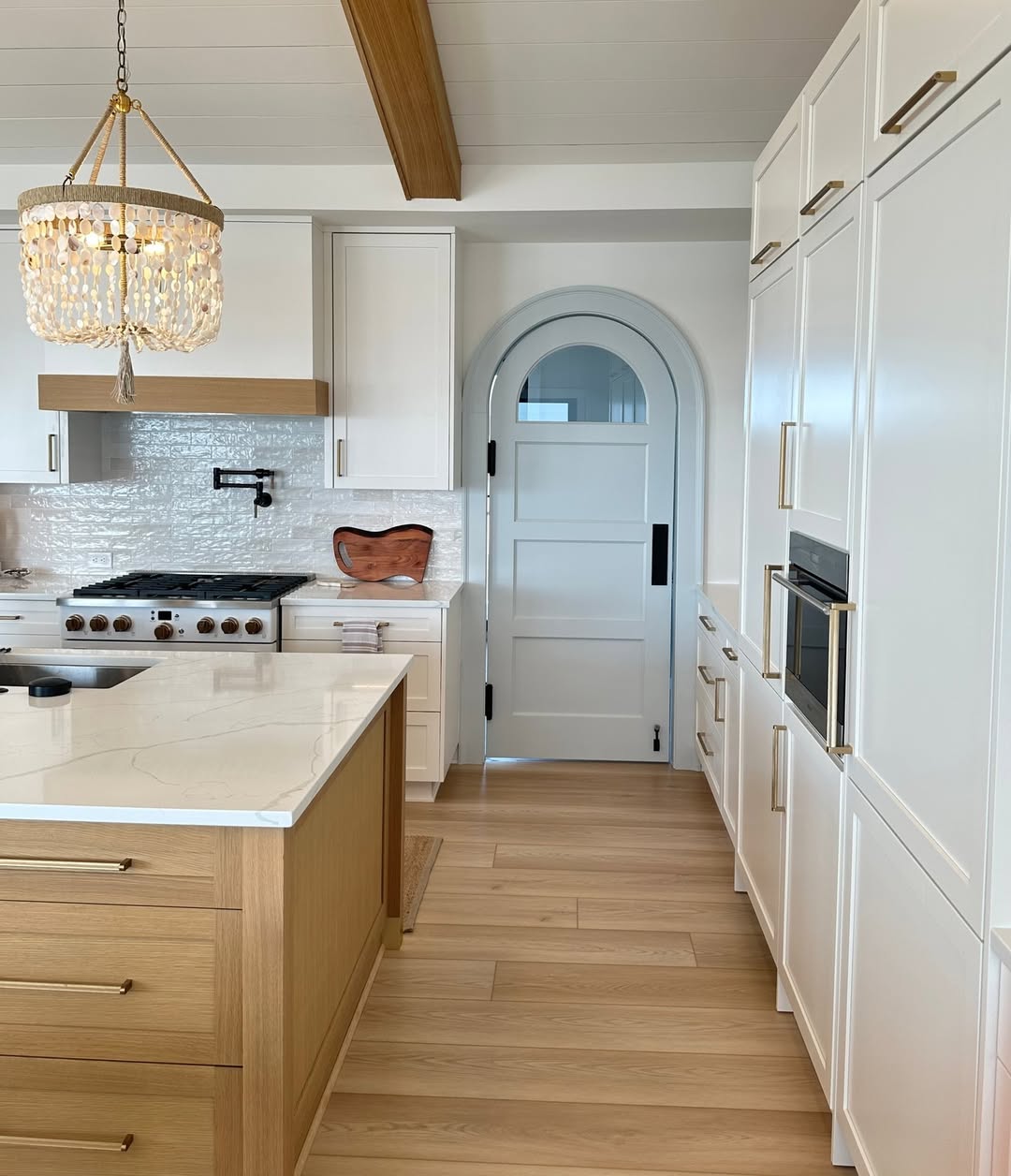 Modern kitchen with white cabinets, wood accents, a central island, stainless steel appliances, and a stylish arched pantry door in the background—a great addition for those seeking inspiring pantry door ideas. A chandelier hangs above the island.