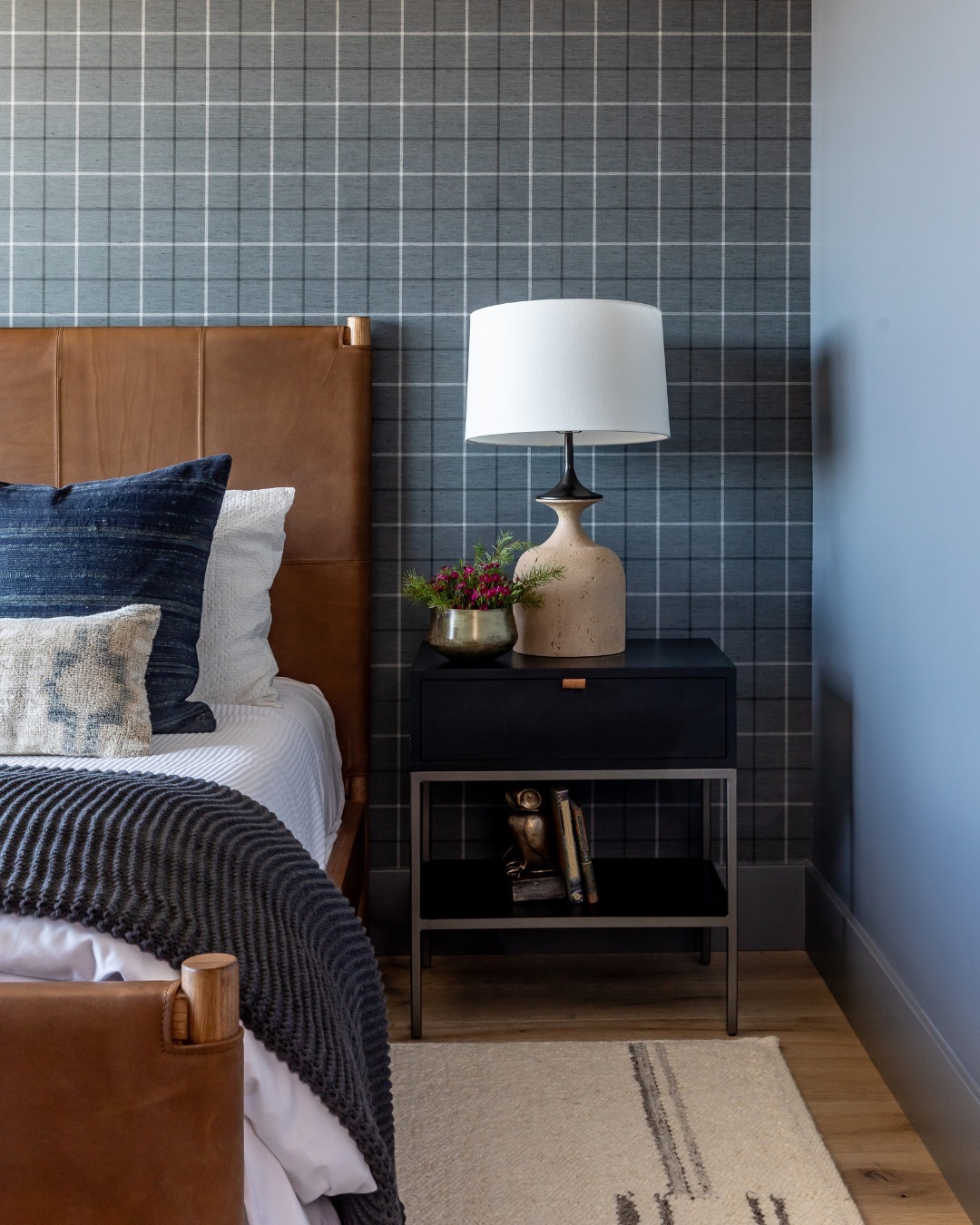 A modern bedroom design featuring a brown leather headboard, navy and white bedding, a black nightstand with a lamp, small plant, and books against a gray plaid accent wall—perfect for stylish bedroom interior ideas.