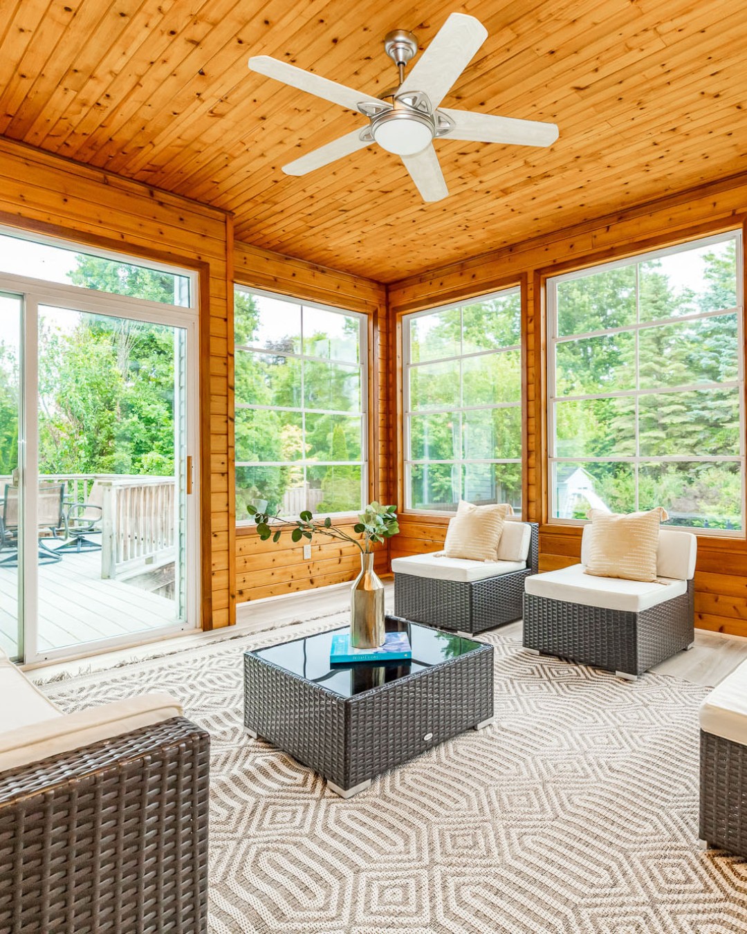 A sunroom designed as a 3 season porch features wood-paneled walls and ceiling, large windows, wicker furniture with cushions, a glass coffee table, and a ceiling fan, overlooking a deck and lush greenery—perfect for porch ideas.