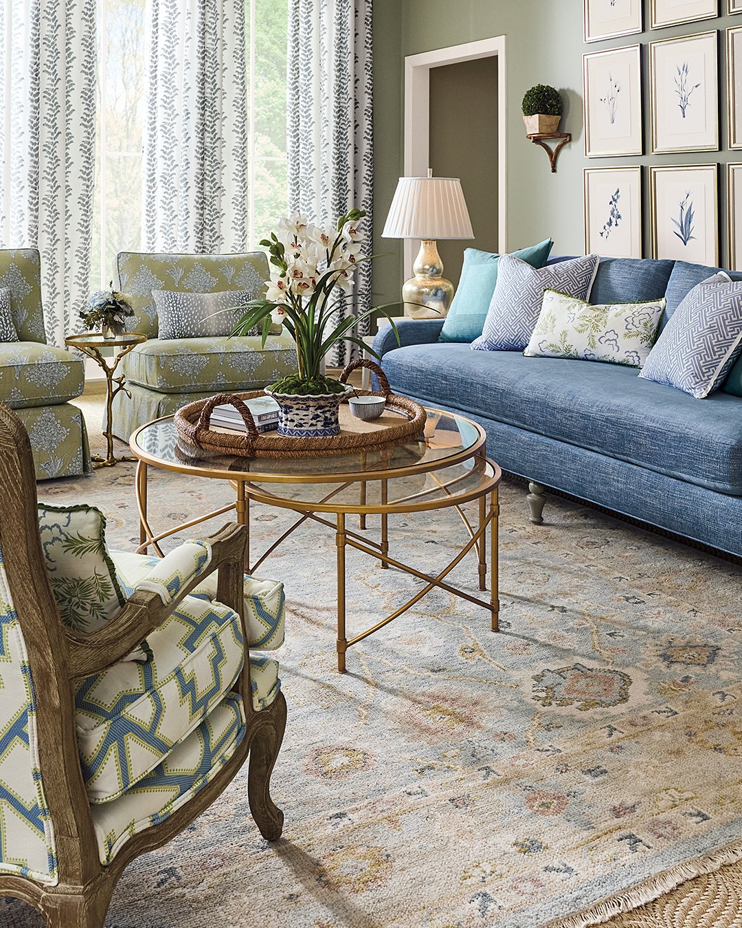 A modern living room design featuring patterned armchairs, a blue sofa, a round glass coffee table, decorative pillows, a soft rug, and wall art with botanical prints creates a stylish and inviting space.