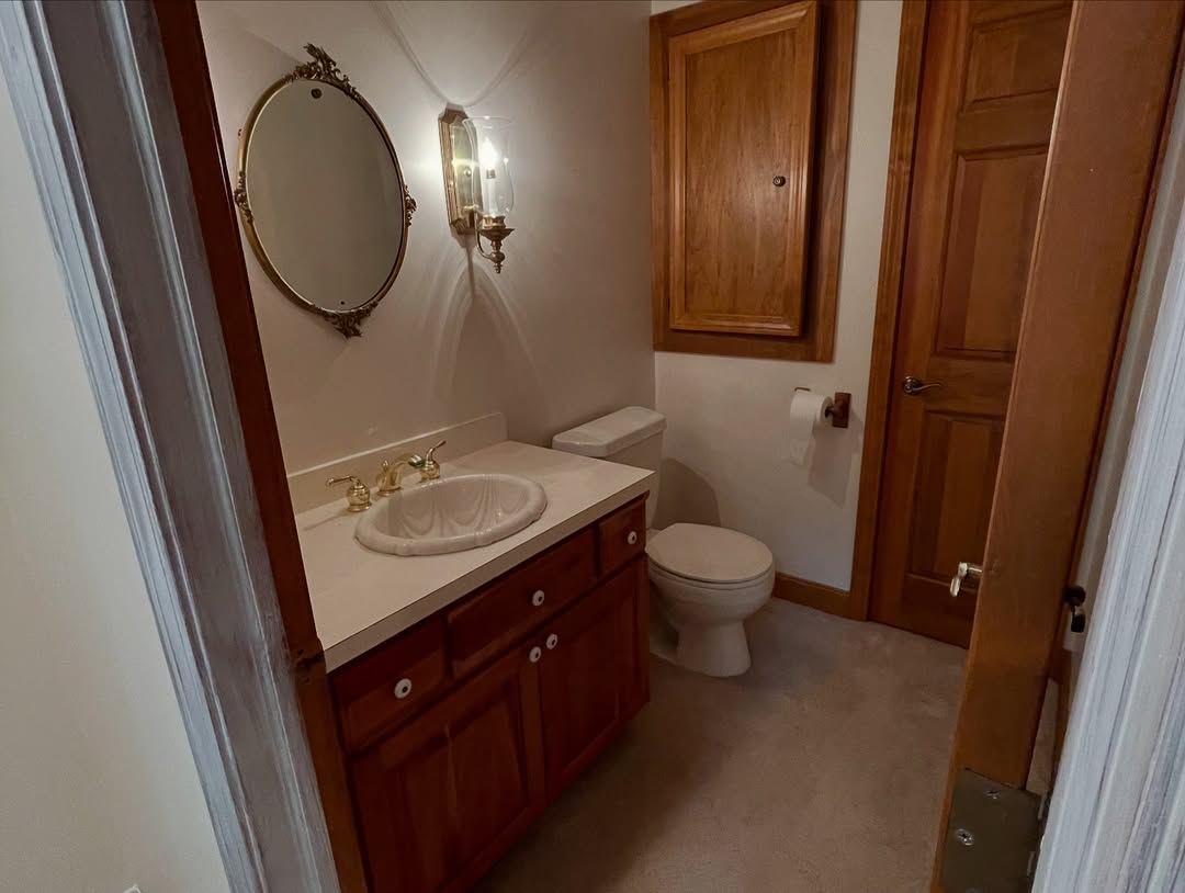Small bathroom with a vanity sink, oval mirror, wall sconce, toilet, wooden cabinetry, and a closed wooden door—perfect for stylish powder rooms that deliver wow-factor in compact spaces.