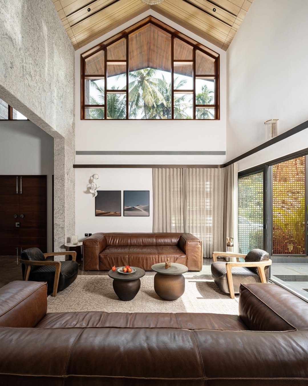 A modern high ceiling living room with large windows, brown leather sofas, wooden armchairs, and round black coffee tables. Sunlight streams in, highlighting neutral decor and tropical views—perfect for those seeking high ceiling living room ideas.