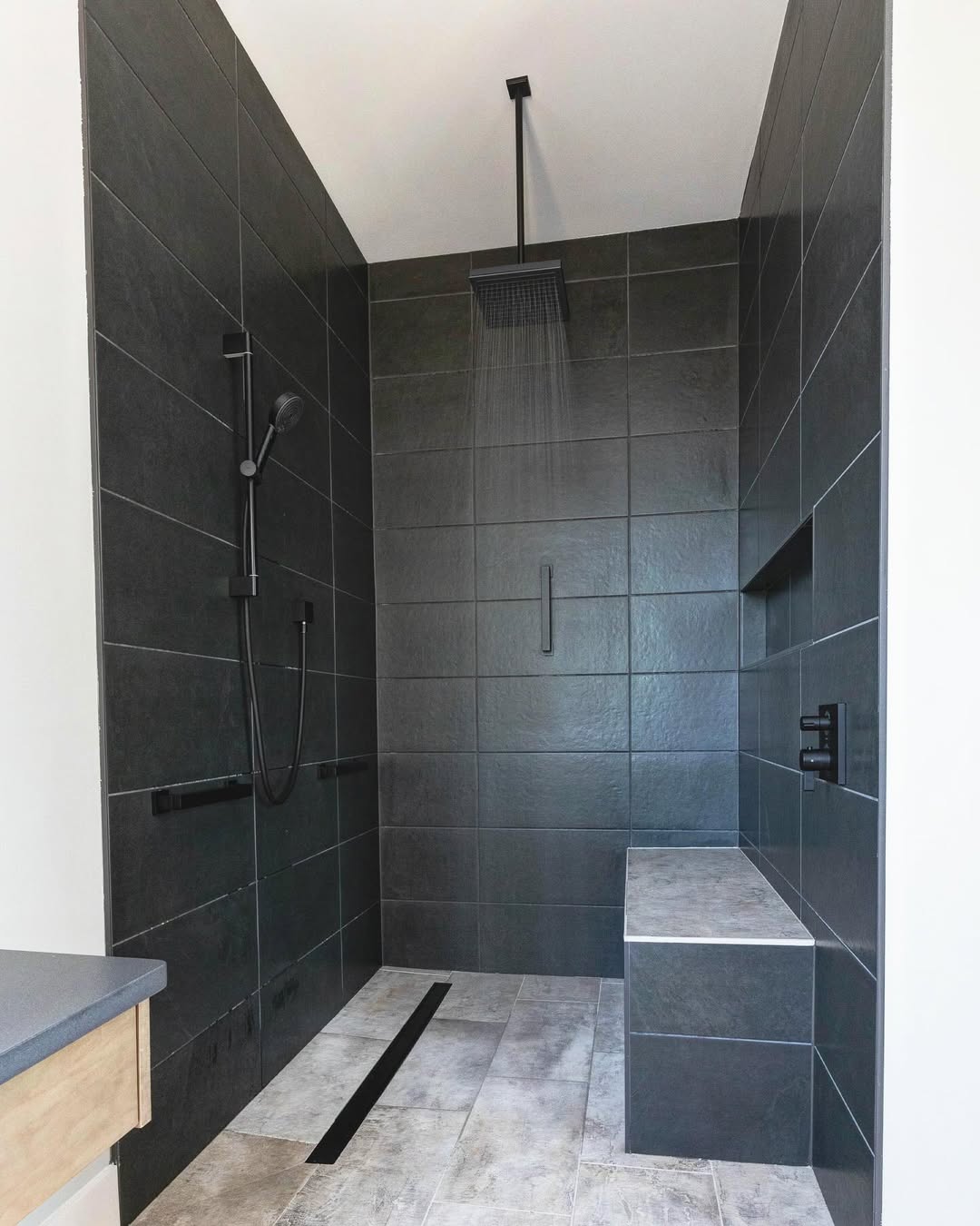 A modern no door shower with dark tiles, a rainfall showerhead, a handheld shower, a built-in bench, and a recessed shelf—an ideal walk-in shower idea for small bathrooms.