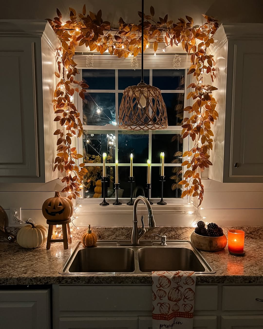 A kitchen sink area with autumn leaf garland and fairy lights framing the window, lit candles, a jack-o’-lantern, pumpkins, and a candle on the counter—perfect Halloween Kitchen Decor for cozy fall vibes.