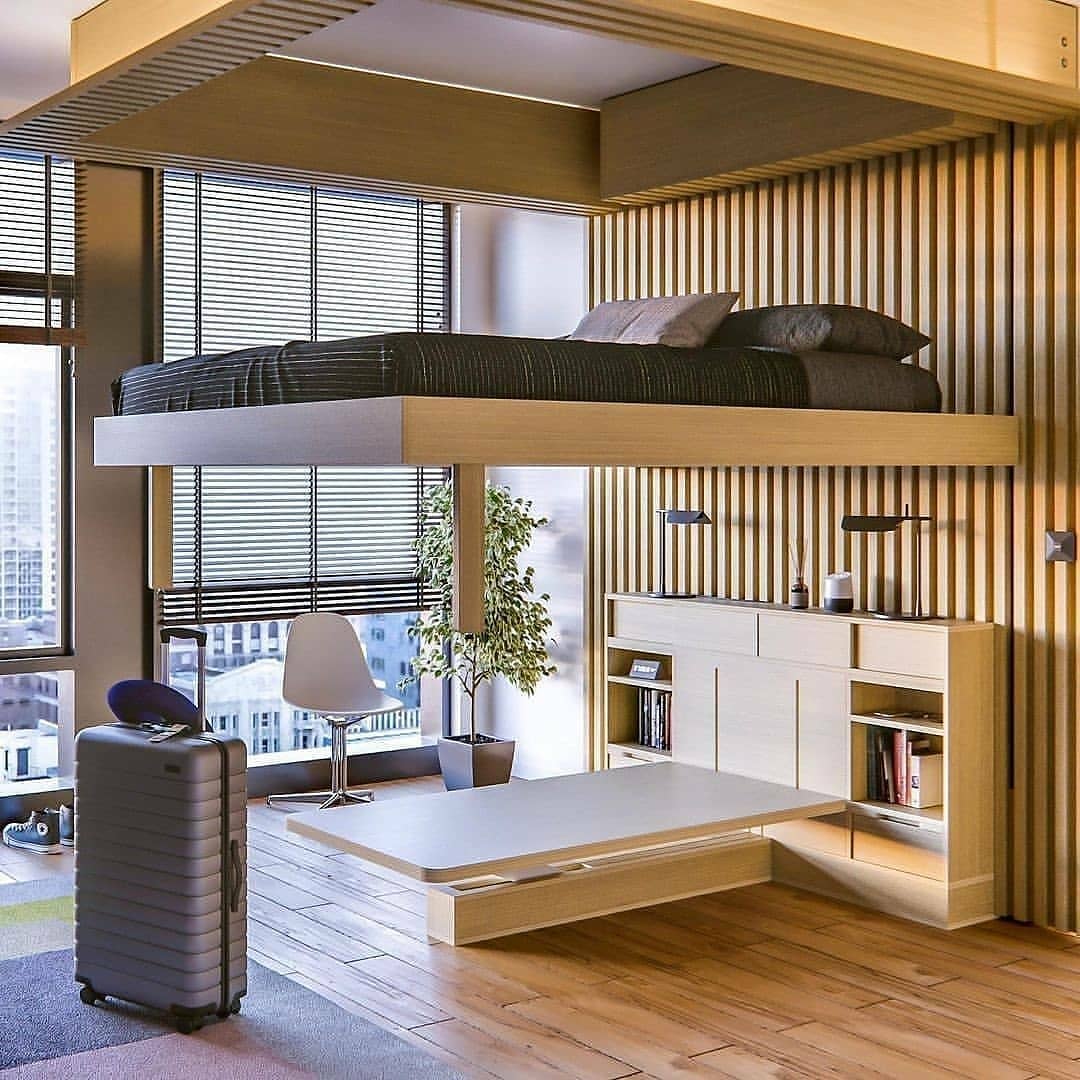 A modern apartment featuring space saving furniture: a suspended bed above a built-in desk, cabinets, and shelves; a suitcase and chair are nearby, framed by large windows in the background—smart furniture ideas for maximizing space.