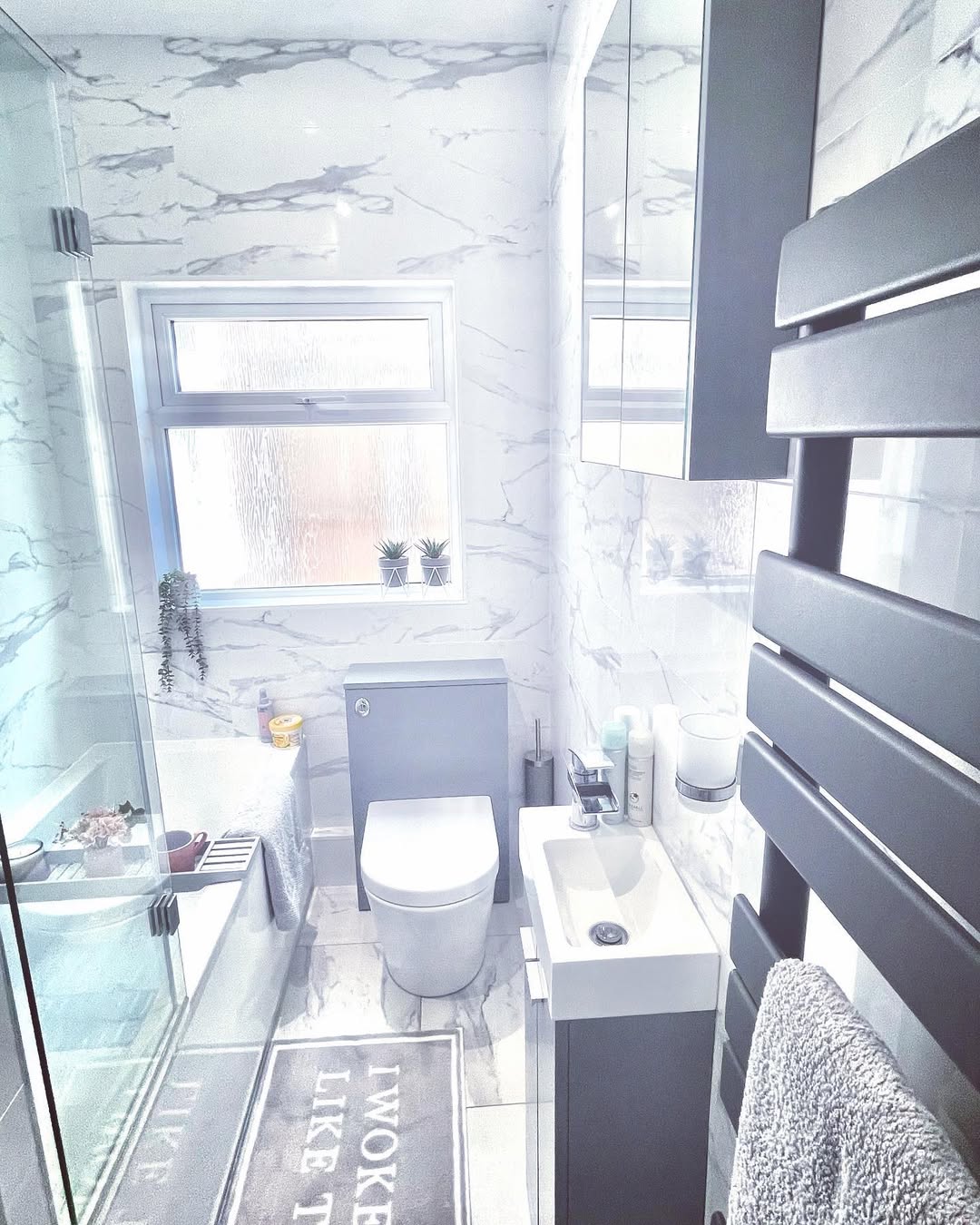 Modern bathroom with white marble walls, a space saving bathtub with glass screen, grey vanity, wall mirror cabinets, towel radiator, and a window; two small plants sit on the sill—perfect for those seeking furniture ideas.