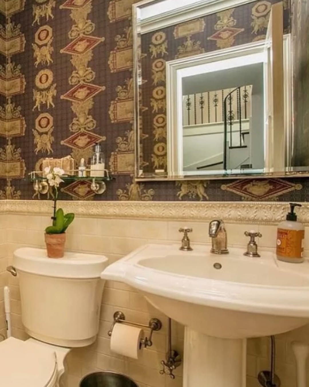 A stylish powder room with patterned wallpaper, a pedestal sink, wall mirror reflecting a staircase, potted orchid, and chic details—perfect for wow-factor powder room ideas.
