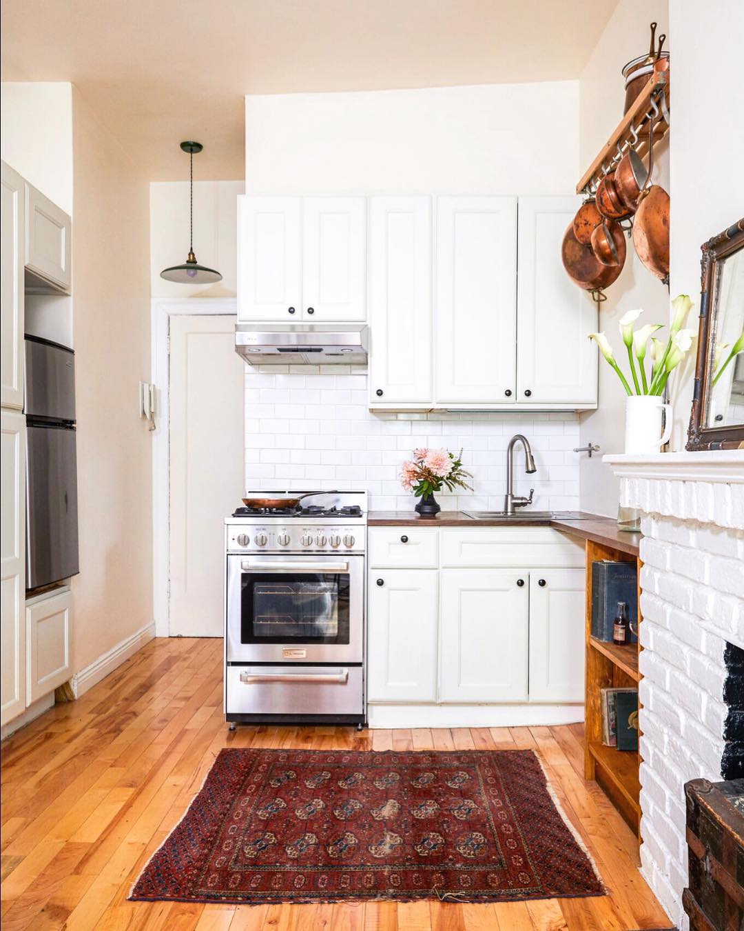 A small kitchen with white cabinets, a stainless steel stove, a red patterned rug, wooden floors, and hanging copper pots showcases smart apartment organization and charming small space decor with flowers in vases on the counter and mantle.
