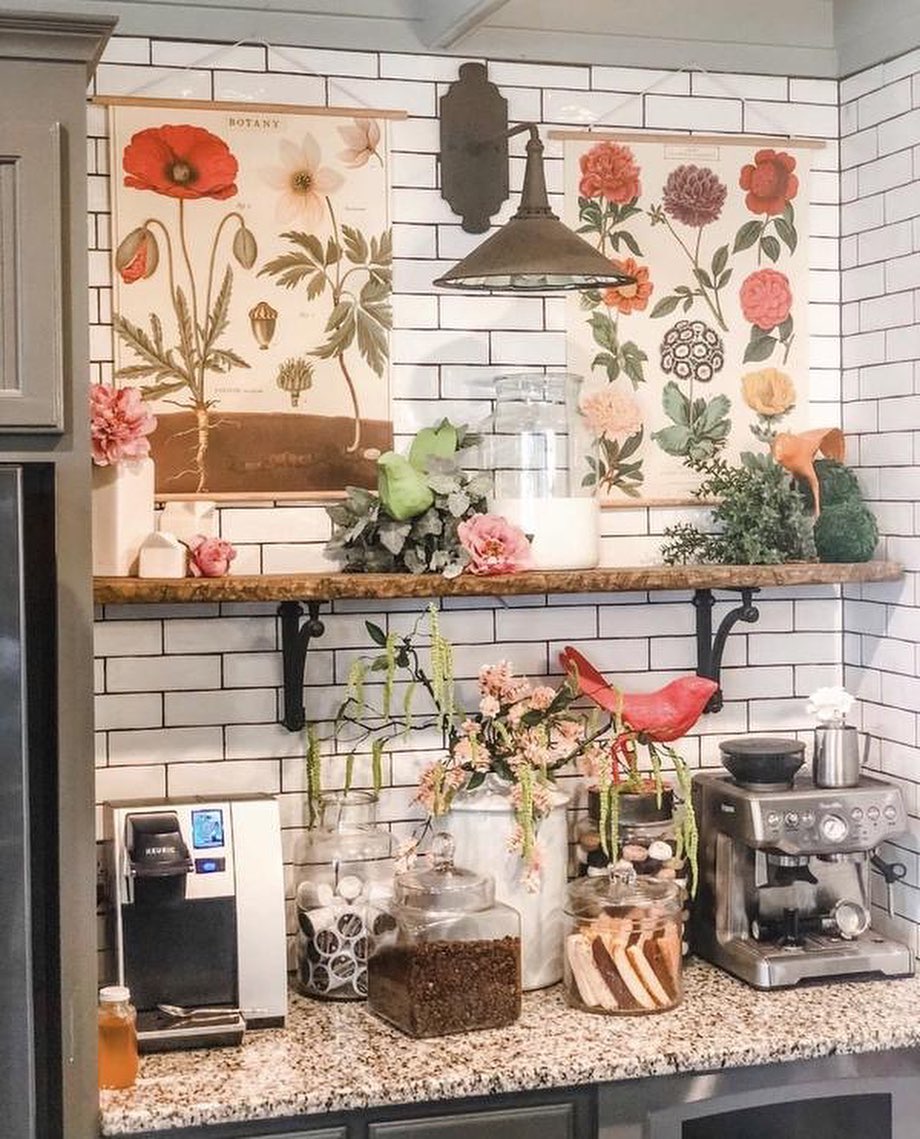A kitchen corner transforms into a cozy coffee bar, featuring a coffee machine, jars, and mugs on the countertop, with floral decor, plants, and botanical prints brightening the white subway tile backsplash—perfect for kitchen coffee bar ideas.