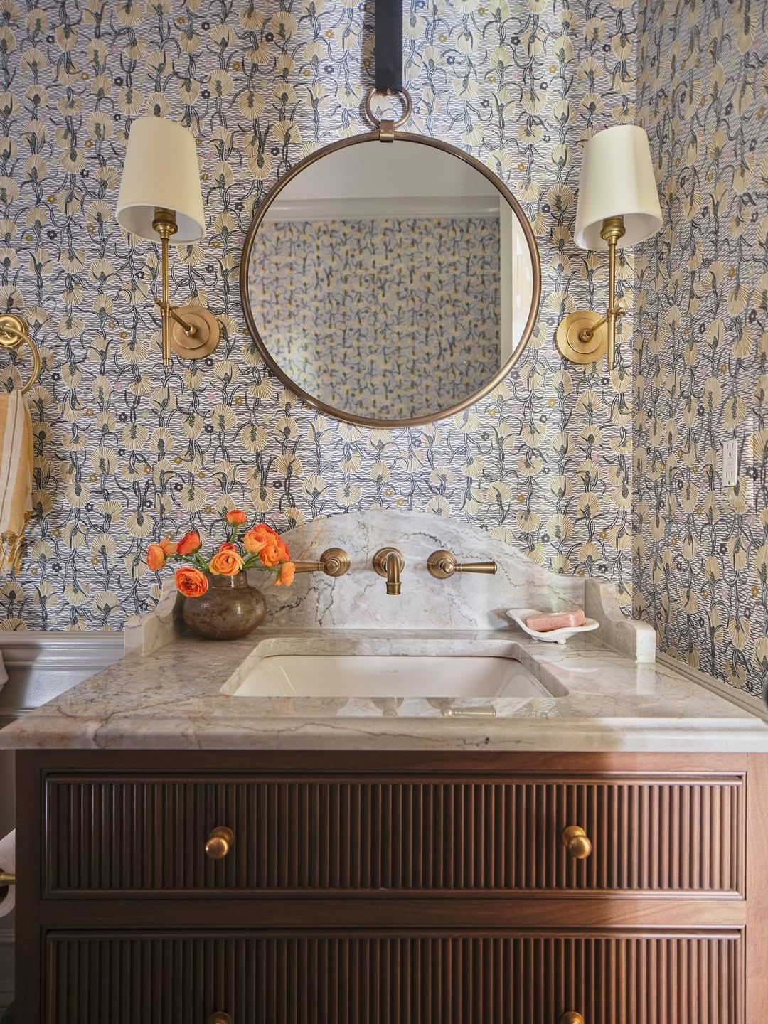 A bathroom vanity with a marble countertop, brass fixtures, round mirror, two wall sconces, floral wallpaper, and a vase of orange flowers creates wow-factor decor—perfect inspiration for stylish powder rooms.