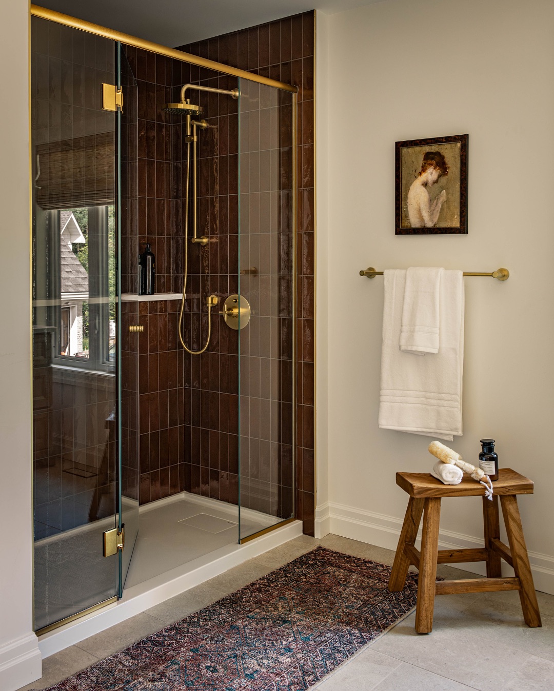 Modern bathroom with a no door walk-in shower featuring gold fixtures, brown tile walls, a wooden stool with toiletries, a towel rack, and a framed portrait—perfect inspiration for small bathrooms.