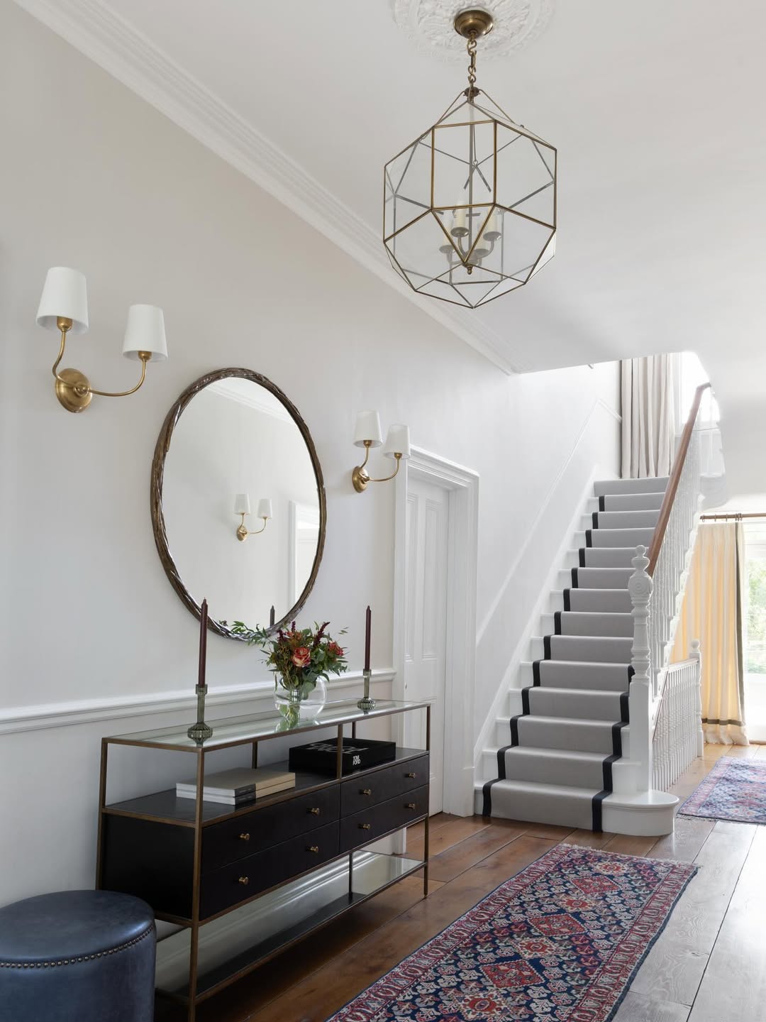 A hallway with a console table, round mirror, two wall sconces, and a runner rug showcases timeless hallway design. A staircase with a gray carpet runner leads upstairs as natural light enters from the far end.