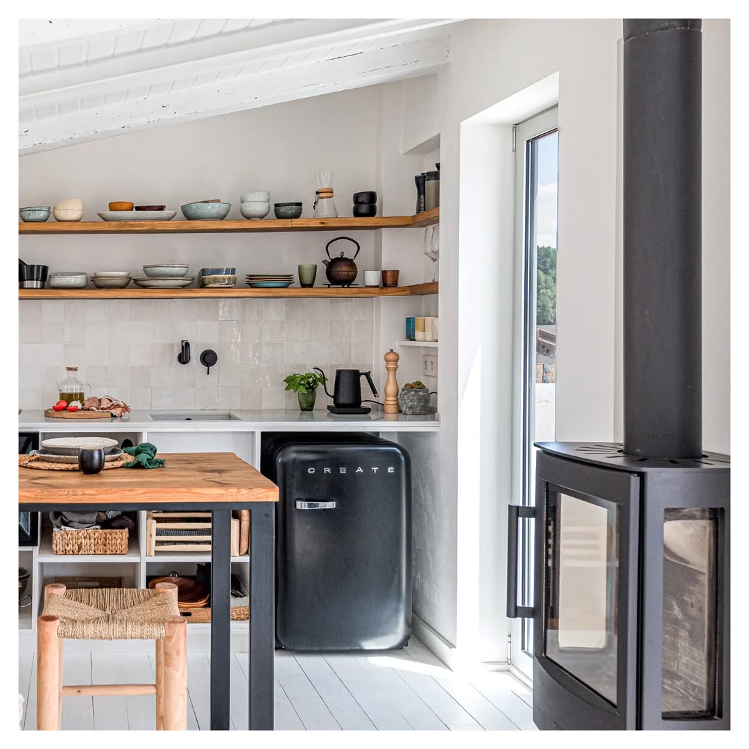 A modern kitchen perfect for small space decorating, featuring open wooden shelves, a black retro fridge, a wood-burning stove, and natural light streaming through a glass door.