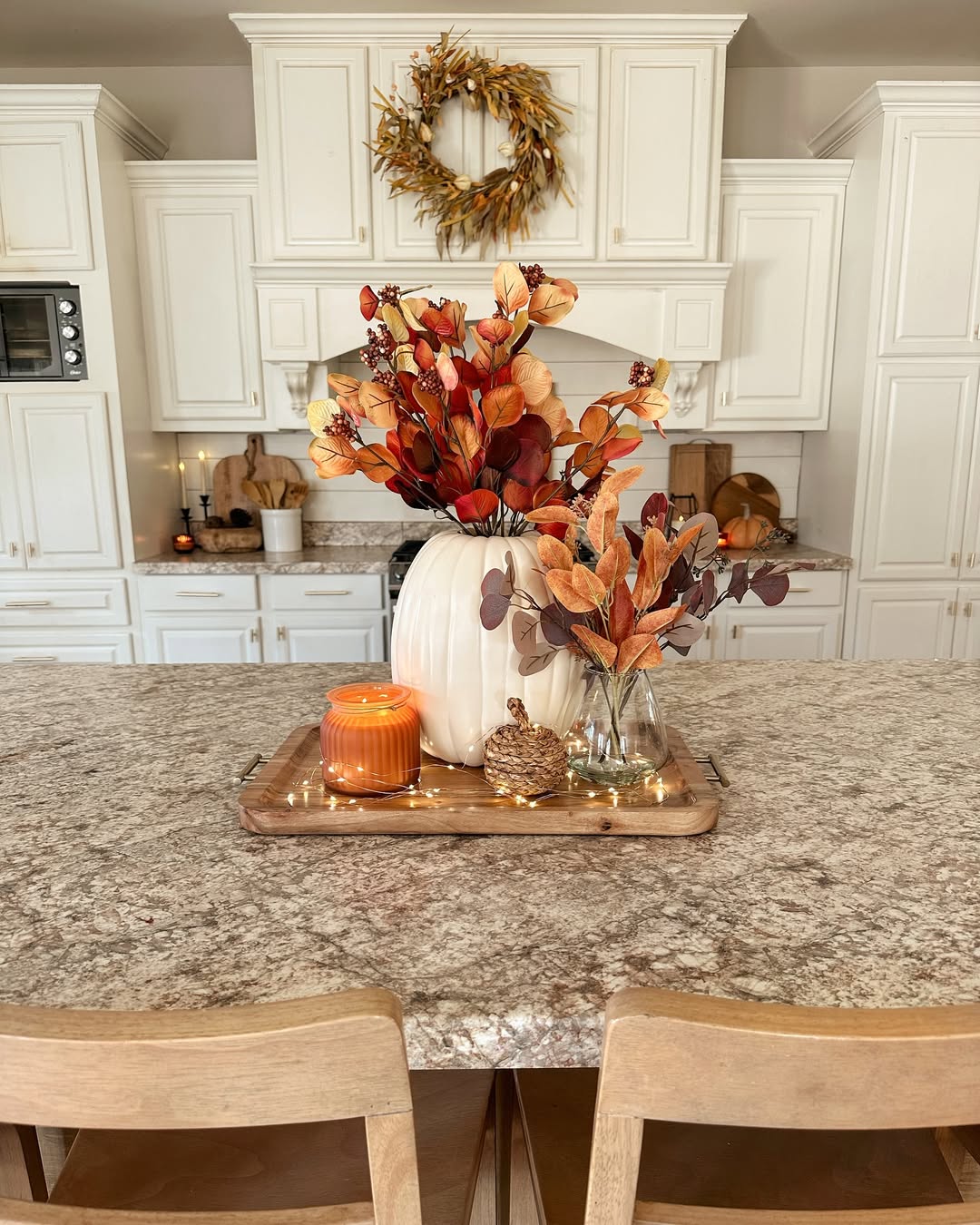 A kitchen island displays autumn-themed decor, including a white pumpkin vase with orange leaves, a small glass vase, an orange candle, and string lights on a wooden tray—perfect inspiration for Halloween decor ideas.