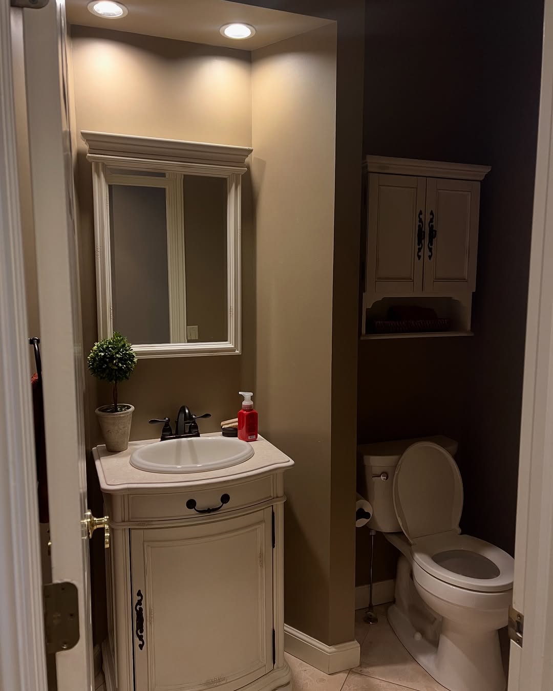 Small bathroom with a white vanity and sink, mirror above, soap dispenser, potted plant, toilet with wall cabinet above, and beige walls—perfect for those seeking wow-factor or stylish powder rooms inspiration.