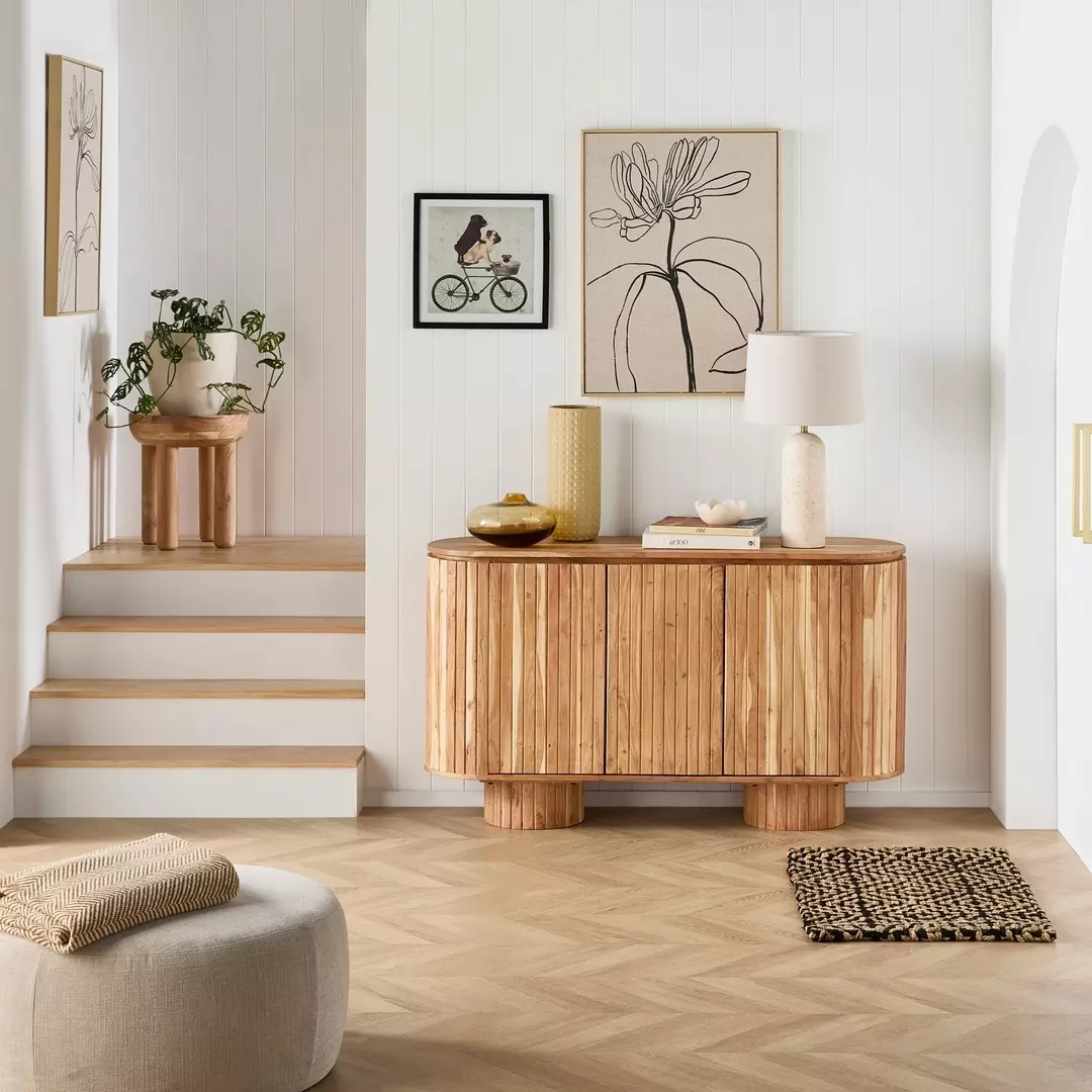 A modern living space with light wood furniture, abstract art on white walls, a small plant, a lamp, and a woven rug on herringbone flooring—perfect for fresh sideboard decor and creative decor ideas.