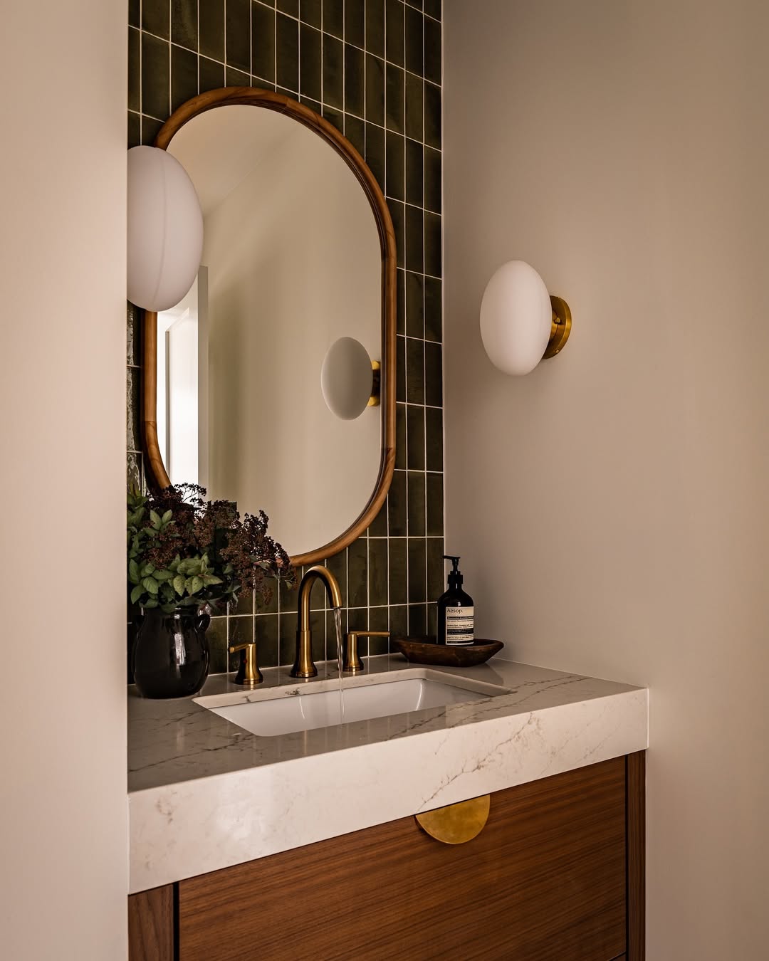 A bathroom vanity with a marble countertop, gold faucet, oval mirror, wall-mounted light, green tile backsplash, and a black vase with flowers—perfect wow-factor decor for stylish powder rooms.