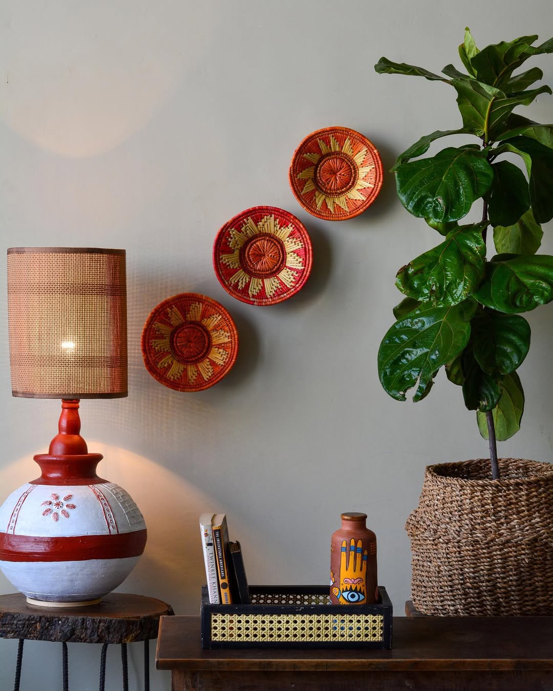 A lamp, a woven basket with a plant, three decorative wall plates—a perfect blend of simple wall decor ideas—plus a table with a bottle and tray, all arranged against a plain wall.