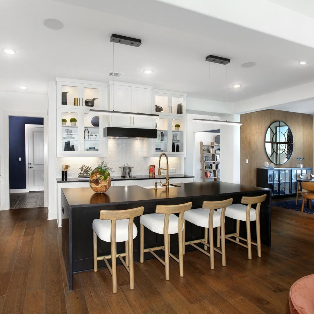 Modern kitchen featuring a black countertop kitchen island, four wooden stools, white cabinets, open shelves, and wood flooring. A round wall clock and console are visible in the adjacent room.