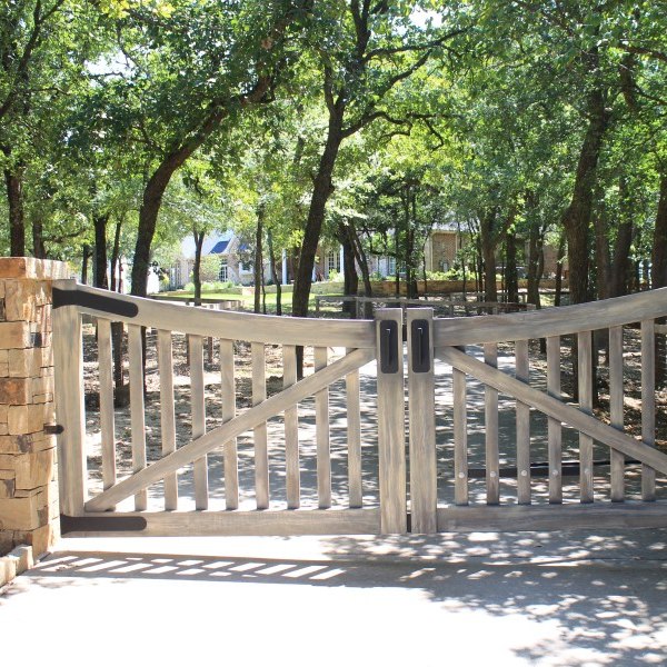 A farmhouse gate blocks a driveway leading to a house surrounded by trees, with sunlight gently filtering through the foliage.