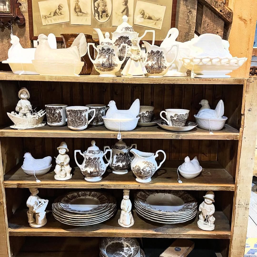 A wooden shelf displays ceramic dishware and figurines, including teapots, bowls, plates, cups, and animal-shaped pieces—perfect inspiration for vintage farmhouse decor—with framed sketches on the wall behind.