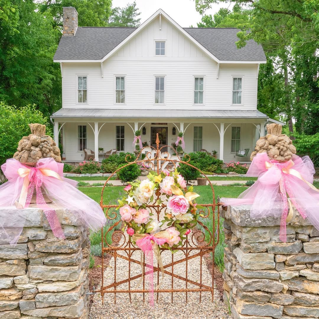 A white two-story house with a large porch sits behind a rusted farmhouse gate adorned with pink ribbons and flowers, flanked by stone pillars and lush greenery.
