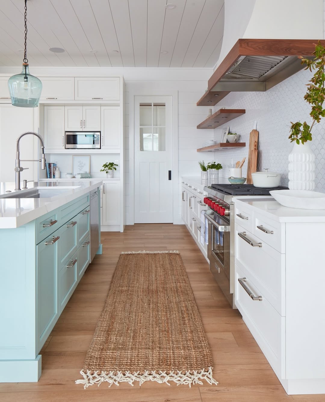 Modern kitchen with white cabinets, a light blue island, stainless steel appliances, open wood shelves, a woven rug, potted plants on a light wood floor, and stylish pantry doors that complement the kitchen design.