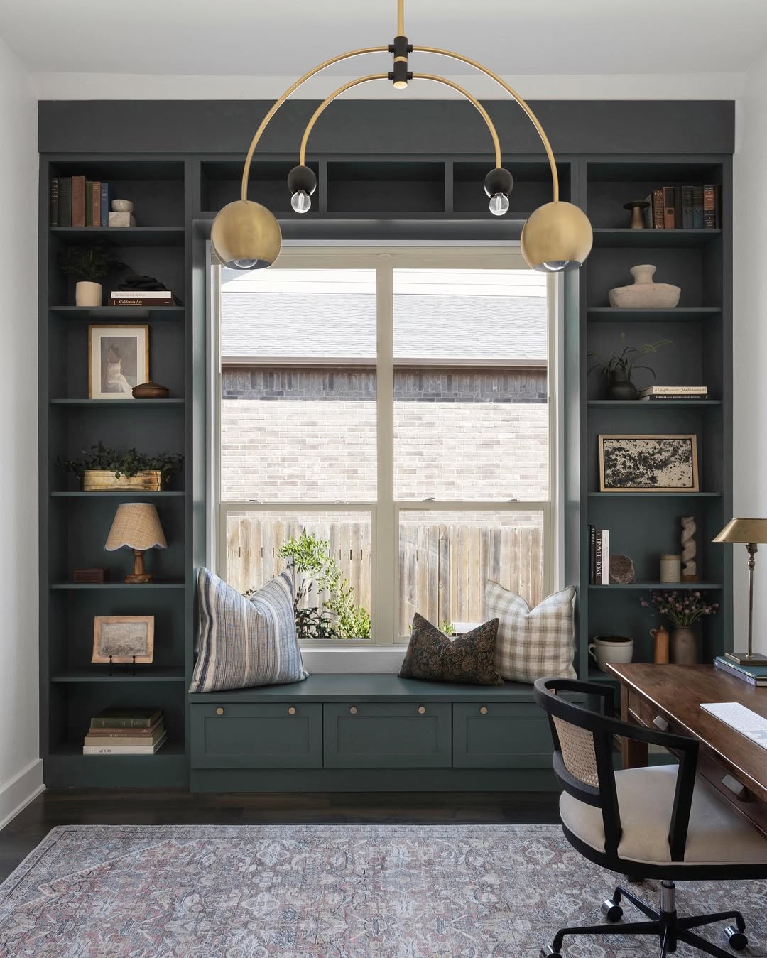 A home office with built-in dark green shelves featuring stylish bookshelf decor, a window seat with cushions, a wooden desk, a modern chandelier, and a patterned rug.