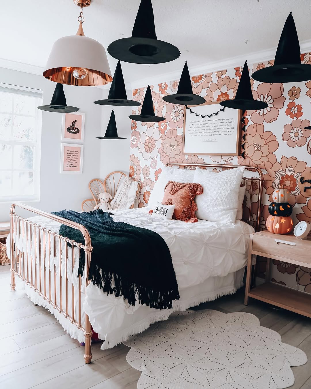 A bedroom with floral wallpaper, a brass bed, white bedding, hanging black witch hats, teddy bears, pumpkin decor, and framed art offers unique Halloween bedroom decor for those seeking creative and spooky bedroom ideas.