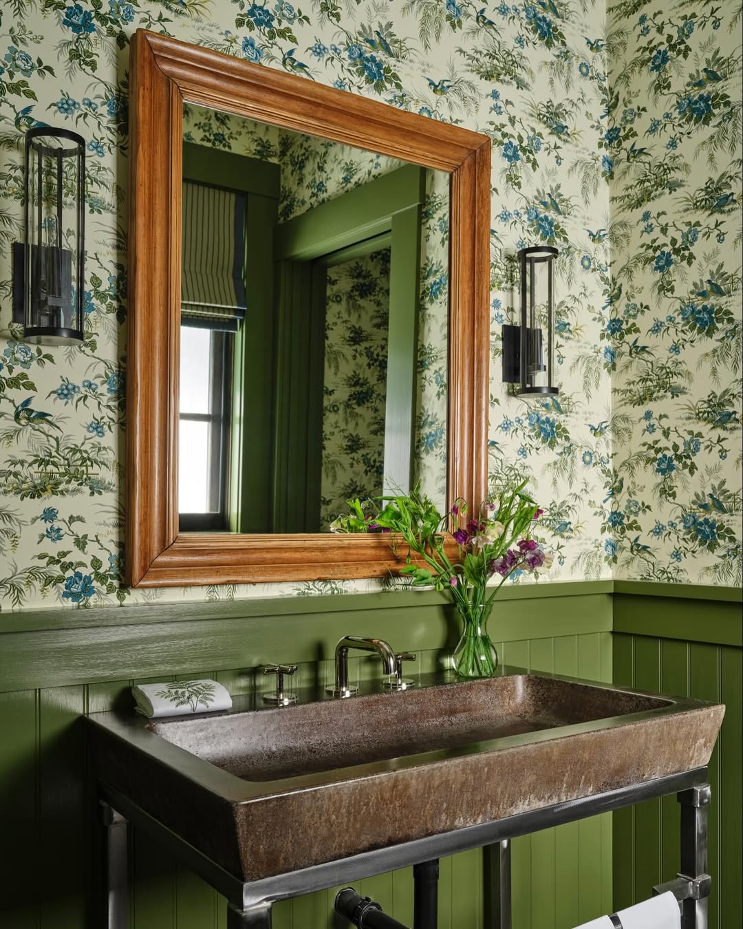 This bathroom showcases wow-factor with floral wallpaper, green wainscoting, a wood-framed mirror, wall sconces, rustic sink, and a vase of flowers—perfect inspiration for stylish powder rooms.