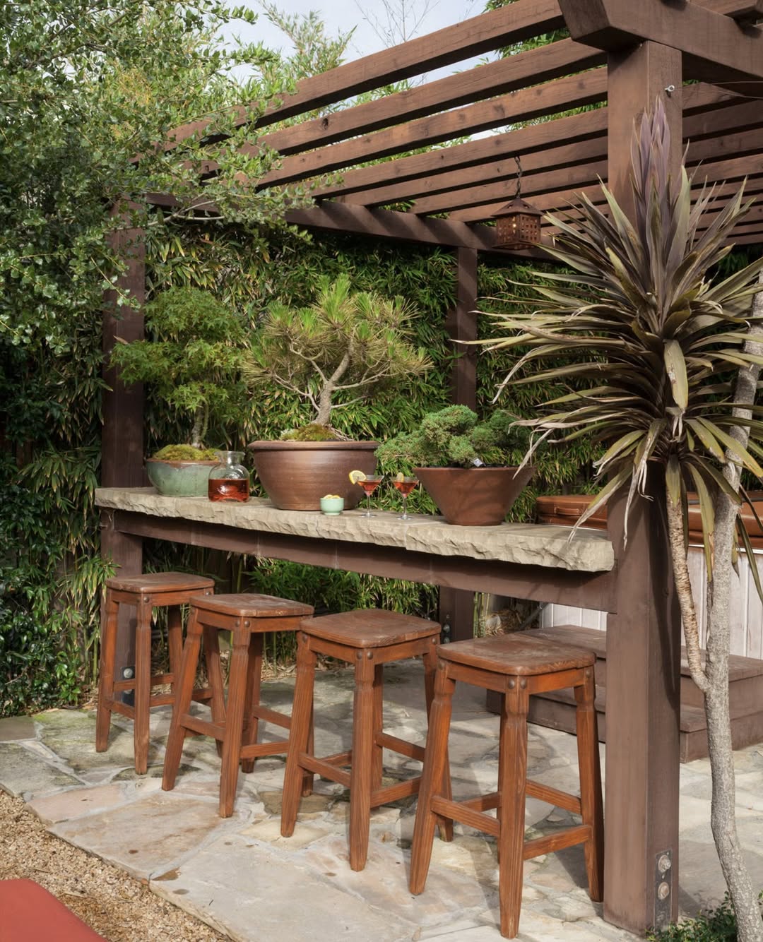 A wooden outdoor bar with four stools, a concrete countertop, and potted plants beneath a pergola surrounded by greenery—perfect inspiration for rustic outdoor kitchen ideas.